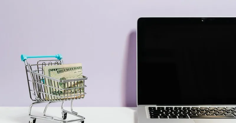 Shopping cart with money next to a laptop symbolizing online shopping and e-commerce.