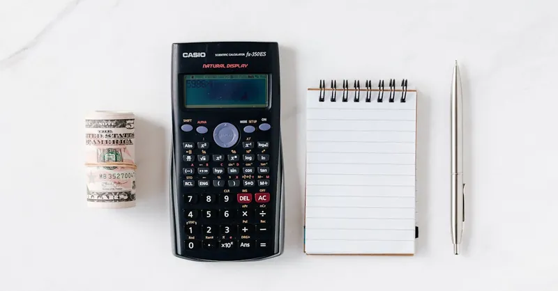 Top view of financial tools including a calculator, notepad, pen, and rolled cash on a white background.