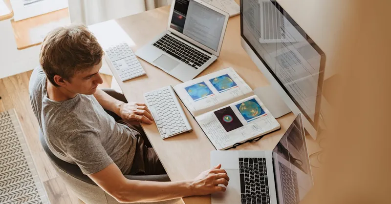 A man multitasks with laptops and a desktop, coding in a home office setting.