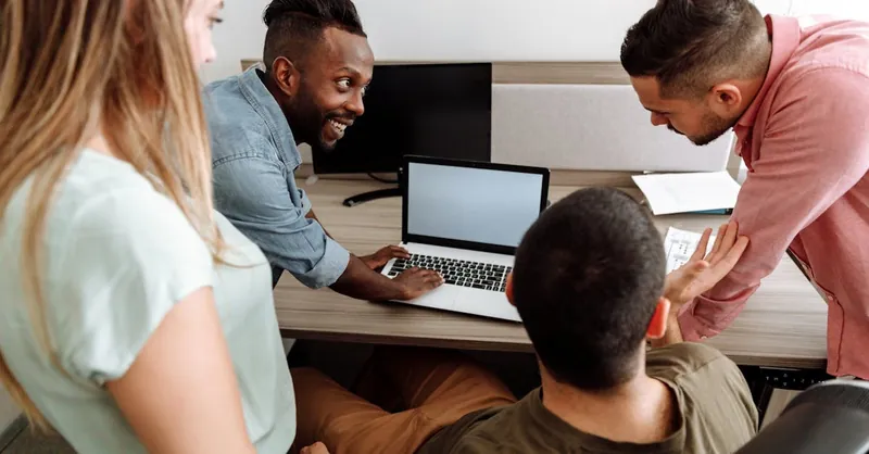 Four colleagues brainstorming around a laptop in a bright, modern office setting.