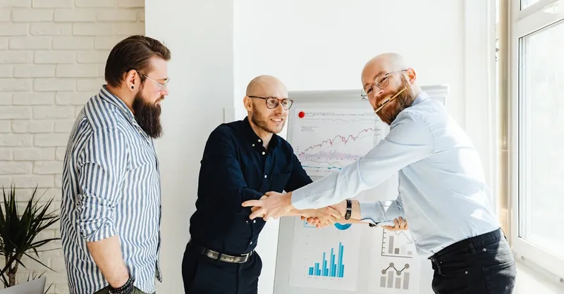 Three businessmen engage in a lively discussion around a whiteboard with charts in a sunlit office.