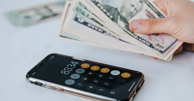 Calculator application on mobile phone screen on white surface near crop anonymous person holding pile of dollars