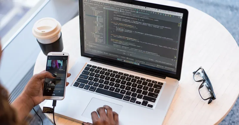 A programmer manages code on a laptop while multitasking with a smartphone, illustrating modern tech workspace.