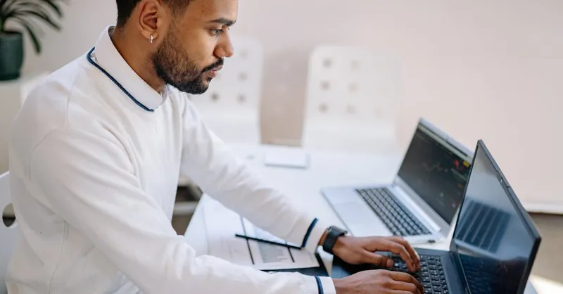 Young professional working diligently on multiple laptops in a modern office setting.