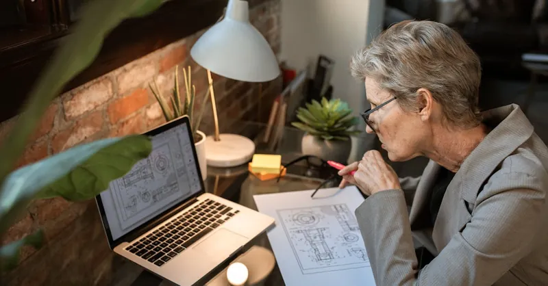 A senior engineer analyzes architectural plans on a laptop in a cozy home office.