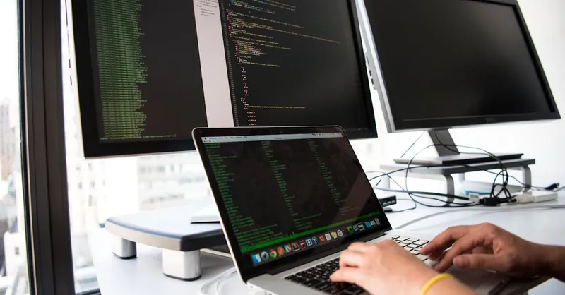 A developer writes code on a laptop in front of multiple monitors in an office setting.